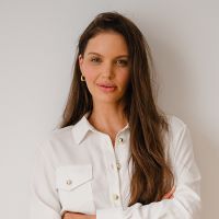 Portrait of a woman with long brown hair wearing a white button-down shirt, standing against a light gray background, arms crossed lightly.