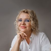 Portrait of a smiling woman with blonde curly hair and glasses, resting her chin on her hand in a white button-down shirt.