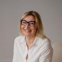 Smiling woman with blond hair and dark glasses wearing a white button-down shirt, seated against a neutral background.